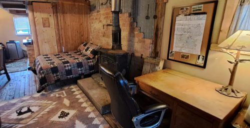 Cozy rustic room with a bed, wood stove, desk, and patterned rug, featuring wooden walls and a map on the wall.